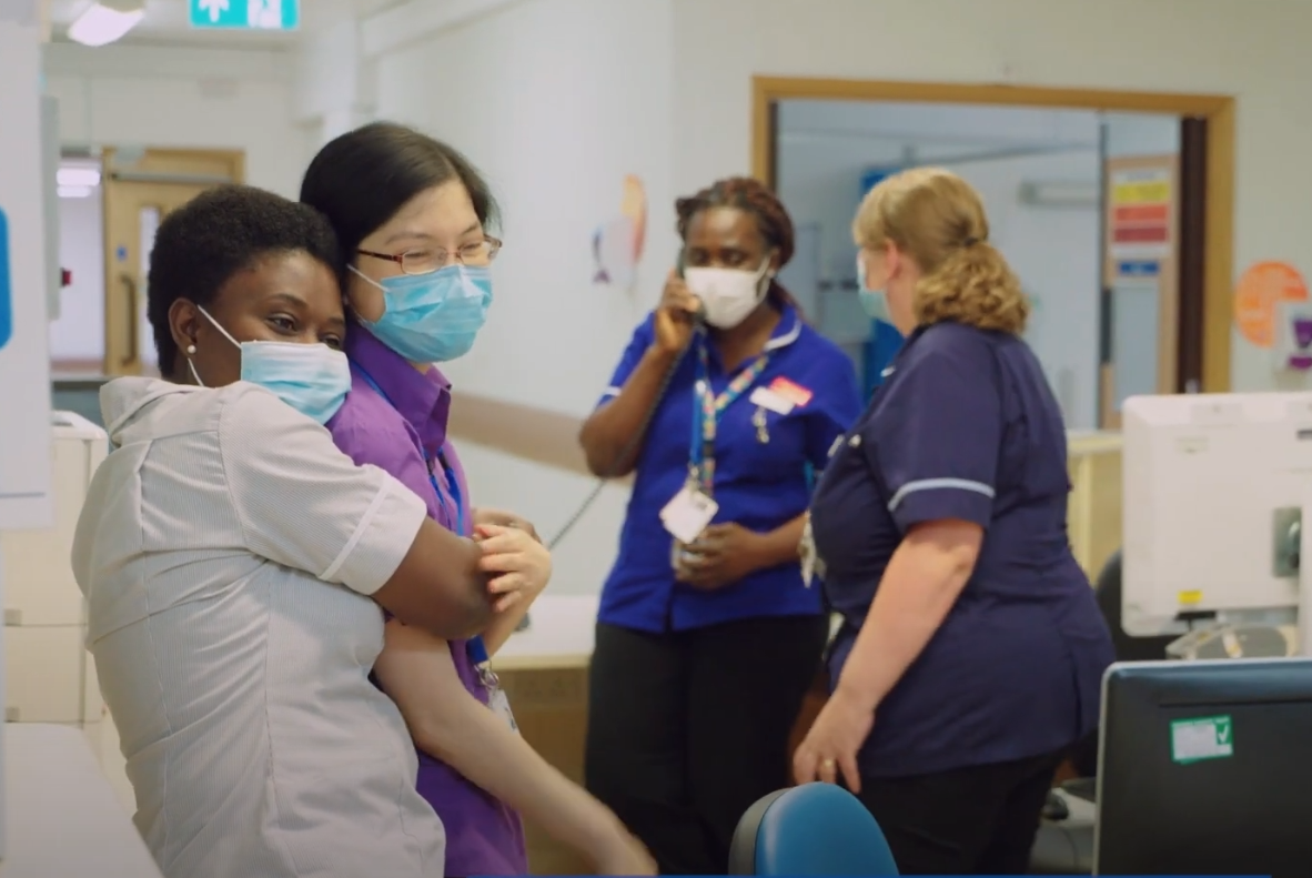 photograph of a nurse hugging a colleague in a busy nurses station and both smiling whilst wearing a face mask